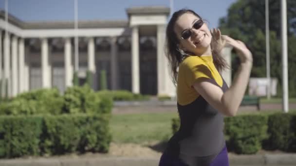 Портрет красивой ретро-женщины, танцующей в солнечном городе на открытом воздухе. Улыбающаяся уверенная кавказка в винтажной спортивной одежде и солнцезащитных очках, наслаждающаяся дискотечной музыкой на улице. — стоковое видео