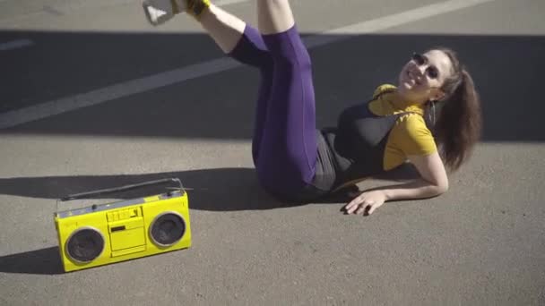 Retrato de jovem alegre em sportswear e óculos de sol sentado na estrada de asfalto e pernas em movimento. positivo caucasiano 1980 esportista posando com retro amarelo gravador de fita na rua de verão. — Vídeo de Stock