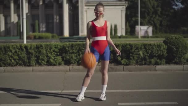 Retrato de mujer joven delgada confiada en ropa deportiva roja posando con pelota de baloncesto al aire libre y sonriendo a la cámara. Hermoso ajuste atleta en gafas de sol
