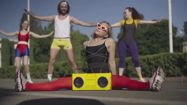 Flexible slim Caucasian sportswoman sitting on twine on road with vintage yellow tape recorder as fit man and women exercising at the background. 1980s sport and healthy lifestyle. — Stock Footage Flexible slim Caucasian sportswoman sitting on twine on road with vintage yellow tape recorder as fit man and women exercising at the background. 1980s sport and healthy lifestyle. Stock Video