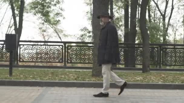 Camera follows careful spy or detective in elegant outfit walking along park alley on cloudy day. Side view wide shot of confident serious Caucasian man in hat and coat strolling outdoors. — Stock Video