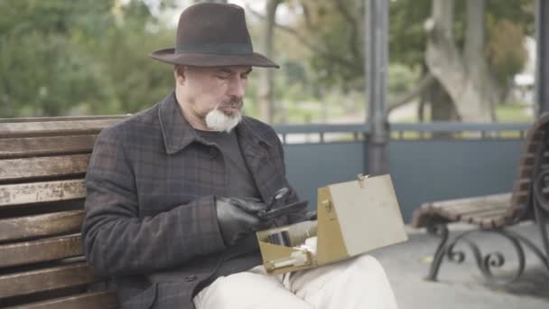 Serious handsome Caucasian spy or intelligencer examining box with novichok using magnifying glass. Portrait of professional man sitting on bench in spring or autumn park with box of toxins. — Stock Video