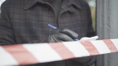 Deri eldivenli tanınmayan bir adam bant bariyerinin arkasında duruyor ve notlar yazıyor. Polis dedektifi ya da istihbarat ajanı bulutlu baharda ya da sonbahar gününde açık havada suç araştırması yapıyor..