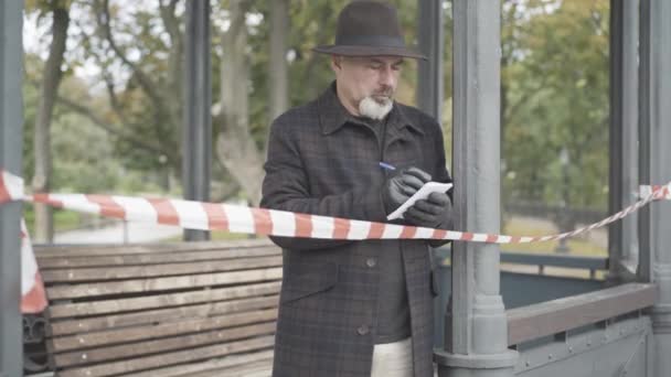 Allvarlig polis detektiv eller underrättelsetjänst står i paviljongen omgiven av byråkrati och vit tejp barriär och skriva anteckningar. Yrkesutredare som utreder brott i vår- eller höstparken — Stockvideo