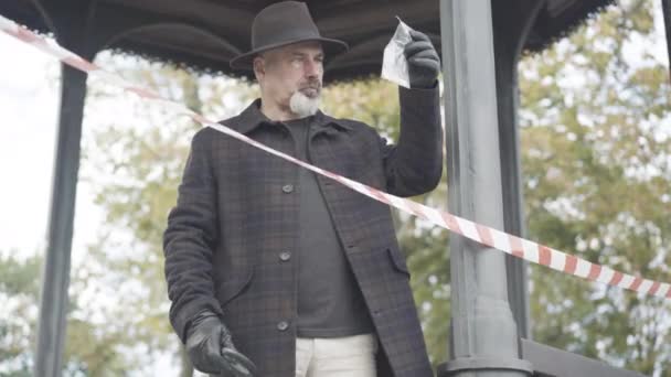 Portrait of thoughtful elegant investigator or detective standing behind red and white tape barrier and examining package with pills using magnifying glass. Professional investigator outdoors. — Stock Video