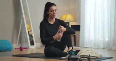 Kendine güvenen zayıf bir kadının evde antrenman yaptıktan sonra şişeyi açıp ferahlatıcı su içtiği geniş bir çekim. Beyaz kadın sporcunun evde egzersiz yapan portresi. Sinema 4k ProRes Merkezi.