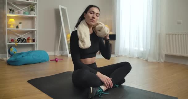 Sportswoman cansado transpirando esfregando o rosto com toalha branca e sorrindo. Retrato de mulher caucasiana magro confiante sentado no tapete de exercício após o treino. Câmara ao vivo. Cinema 4k ProRes HQ. — Vídeo de Stock