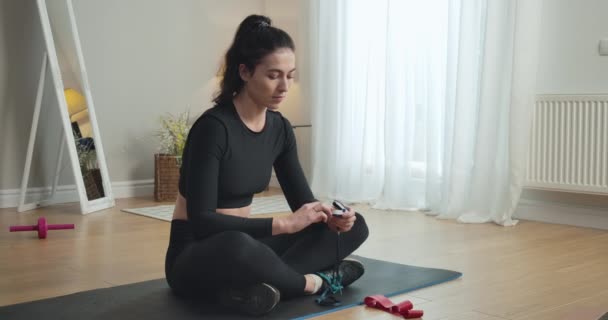 白人黑发女人用脉动血压计测量氧饱和度。健康的年轻女运动员在家锻炼后坐在运动垫上的画像。Cinema 4k ProRes HQ. — 图库视频影像