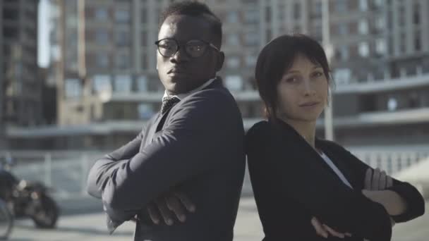 Magabiztos afro-amerikai férfi és kaukázusi nő háttal állnak egymásnak, keresztbe teszik a kezüket és a kamerába néznek. a városi negyedben napfényben pózoló fiatal üzletemberek portréja. Sokszínűség — Stock videók