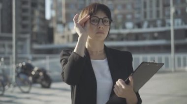 Gözlüklü başarılı kadının portresi güneşli şehir caddesinde duruyor ve kameraya bakıyor. Açık havada poz veren zarif beyaz iş kadını. Güven ve başarı kavramı.