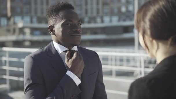 Ung kaukasisk kvinde hjælper afroamerikansk mand med at justere slips og jakke. Portræt af ung forretningsmand stående i sollys med kvinde. Erhvervskoncept. – Stock-video