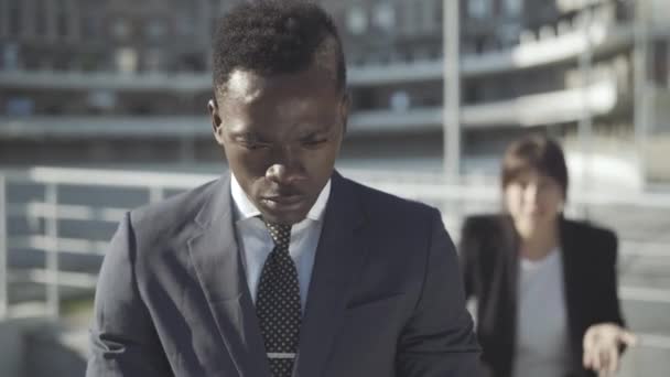 Retrato de un hombre afroamericano molesto parado al aire libre y pensando como una mujer caucásica borrosa gritando y haciendo gestos al fondo. Triste joven empresario que tiene problemas de relación. — Vídeos de Stock