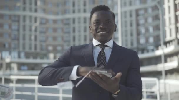 Jeune homme d'affaires prospère dispersant de l'argent et souriant. Portrait d'un homme afro-américain riche profitant d'une journée ensoleillée dans un quartier d'affaires en plein air. Concept de richesse et de succès. — Video