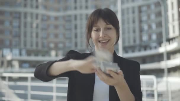 Feliz joven rica mujer dispersando dinero al aire libre y sonriendo a la cámara. Retrato de la acaudalada exitosa empresaria caucásica de pie a la luz del sol en el distrito de negocios. Concepto de riqueza. — Vídeos de Stock