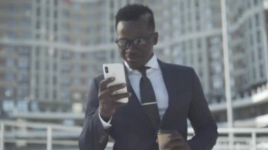Açık havada akıllı telefon kullanarak resmi takım elbiseli ve gözlüklü kendine güvenen Afro-Amerikan bir adam. Başarılı genç işadamının portresi iş bölgesinde internet sörfünde kahve molası veriyor..