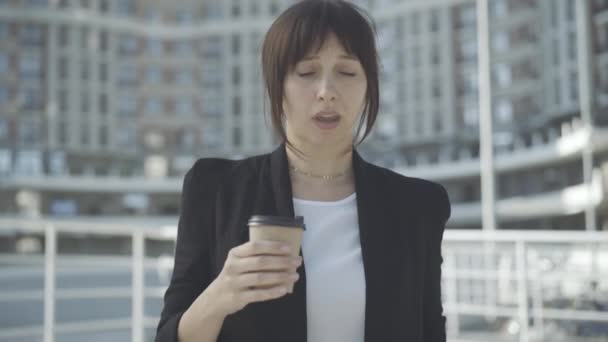 Porträt einer müden jungen Geschäftsfrau, die gähnt und im Freien Kaffee trinkt. Erschöpfte Kaukasierin steht mit Heißgetränk im Geschäftsviertel. Überarbeitetes Konzept. — Stockvideo