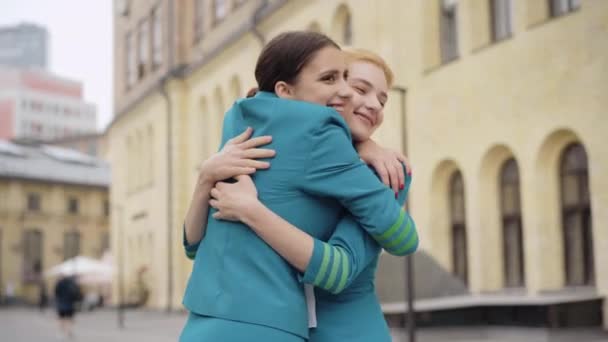 Mellan skott av två smala kvinnor i flygvärdinna uniform kramar på stadens gata. Leende glada unga kaukasiska flygvärdinnor träffas utomhus. Livsstil och turism koncept. — Stockvideo