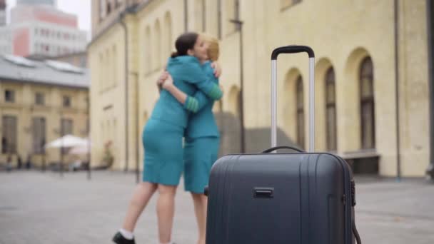 Suitcase standing on city street with two blurred Caucasian women hugging at the background. Wide shot of happy stewardesses meeting after arrival in European town. Elegance and occupation concept. — Stock Video