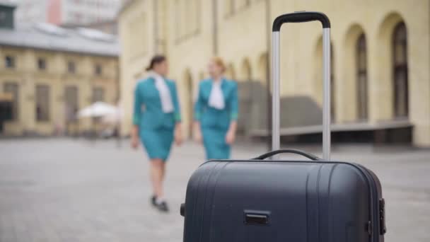Cestovní taška, stojící venku se dvěma rozmazanými štíhlými mladými ženami v uniformě letušky, kráčející do zavazadel po ulici. Profesionální a sebevědomé letušky v evropském městě. Cestovní ruch a zaměstnání. — Stock video
