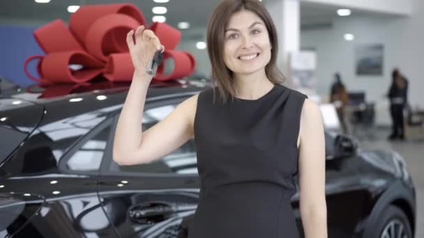 Gelukkig nieuwe eigenaar opscheppen autosleutels in de showroom of dealerovereenkomst. Portret van een glimlachende blanke vrouw die poseert met een zwart voertuig op de achtergrond. Succes en leefstijlconcept. — Stockvideo