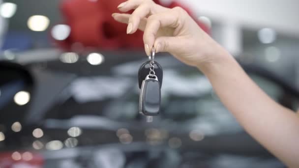 Close-up de mão caucasiana feminino segurando as chaves do carro com o veículo preto embaçado em pé no fundo na concessionária ou showroom. Ireconhecível orgulhoso proprietário comprar automóvel. — Vídeo de Stock