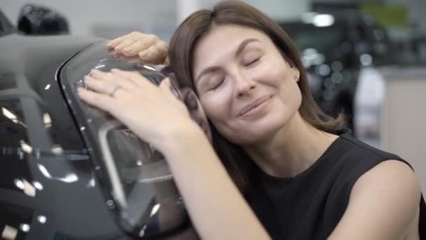 Detailní záběr šťastně se usmívající mladé ženy sedící na černém novém vozidle v prodejně aut. Portrét bohaté brunetky Kavkazské podnikatelky s automobilem v showroomu. Obchod a životní styl. — Stock video