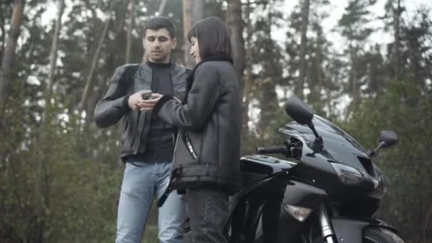 Mladí motorkáři stojící v jarním nebo podzimním lese a odvracející se pohledem dalekohledem. Sebevědomá běloška a muž z Blízkého východu si užívají zatažený den venku. Mezirasový vztah. — Stock video