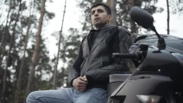 Porträtt av självsäker ung Mellanöstern man står på motorcykel och tänker. Snygg kille i skinnjacka njuter av ensamhet utomhus på mulen mulen dag. Livsstilskoncept. — Stockvideo