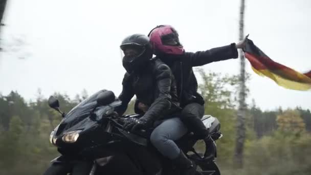 Side view of confident young couple of bikers riding motorcycle holding German flag. Carefree man and woman driving motorbike on cloudy spring or autumn day with Germany national symbol. — Stock Video
