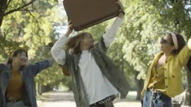 Güneşli yaz yolunda bavul ve ukulele ile dans eden üç kaygısız gülümseyen hippi. 1960 'larda açık havada eğlenen beyaz erkek ve kadınların portresi. Seyahat ve karşı kültür.