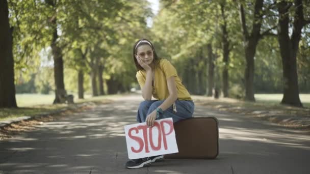 Znudzona hipisowska retro kobieta siedząca na walizce z transparentem Stop. Portret młodej białej kobiety stojącej autostopem na słonecznej letniej drodze w latach 60-tych. Koncepcja stylu życia i podróży. — Wideo stockowe
