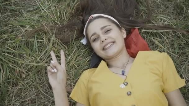 Beautiful young hippie woman lying on green meadow showing peace gesture. Portrait of relaxed carefree Caucasian lady looking at camera and smiling. Counterculture of 1960s. — Stock Video