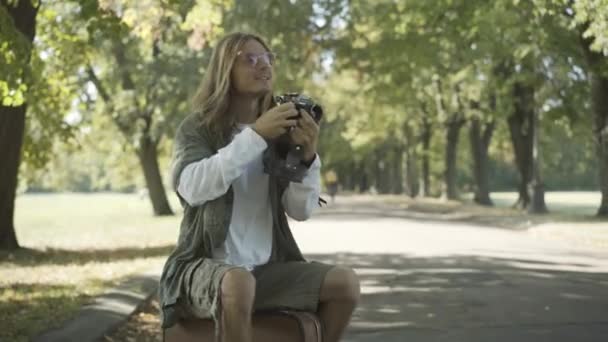 Camera approaches to handsome carefree hippie man sitting on suitcase and taking photos. Positive relaxed Caucasian 1960s guy enjoying travel in sunlight outdoors. Counterculture lifestyle. — Stock Video