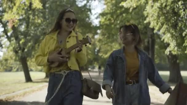 Portret dwóch zrelaksowanych, radosnych hipisek w okularach przeciwsłonecznych spacerujących na świeżym powietrzu w słońcu, grających na ukulele i śpiewających. Śmiejące się kobiety rasy kaukaskiej korzystające z wypoczynku na świeżym powietrzu w latach 60-tych. — Wideo stockowe