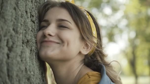 Close-up face of young Caucasian hippie woman hugging tree trunk outdoors. Portrait of carefree beautiful lady enjoying nature on sunny summer day. Lifestyle and 1960s counterculture. — Stock Video