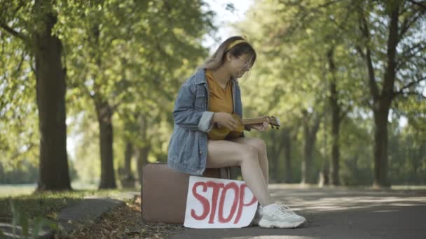 Amplia toma de mujer hippie positiva sentada en la maleta con señal de stop, tocando ukelele y cantando. Retrato de una señora caucásica relajada disfrutando de viajar al aire libre. Autostop y contracultura. — Vídeos de Stock