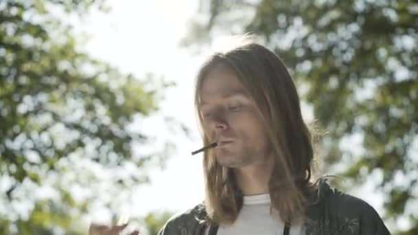 Young Caucasian man lighting cigarette in sunlight outdoors. Portrait of relaxed male hippie smoking in summer park. Concept of 1960s lifestyle and counterculture. — Stock Video