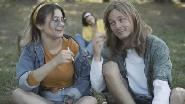Avslappnade par kaukasiska hippies som delar cigaretter utomhus. Porträtt av avslappnad man och kvinna rökning med suddig vän sitter i bakgrunden. 1960-talets retrolivsstil. — Stockvideo