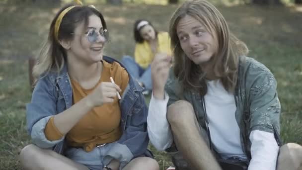 Pareja relajada de hippies caucásicos compartiendo cigarrillos al aire libre. Retrato de un hombre y una mujer relajados fumando con un amigo borroso sentado al fondo. Estilo de vida retro de 1960. — Vídeo de stock