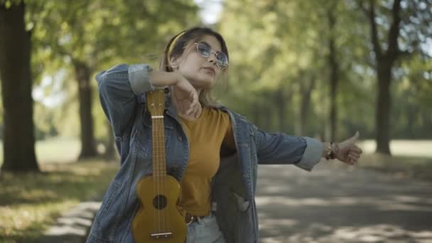 Portrait of young Caucasian woman in sunglasses hitchhiking on sunny summer road. Relaxed female hippie travelling with ukulele outdoors. Tourism and counterculture lifestyle concept. — Stock Video