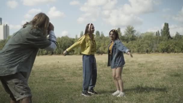 Wide shot of positive hippie women jumping up as man photographing them at camera. Joyful Caucasian friends having fun outdoors on sunny summer day. Counterculture of 1960s concept. — Stock Video