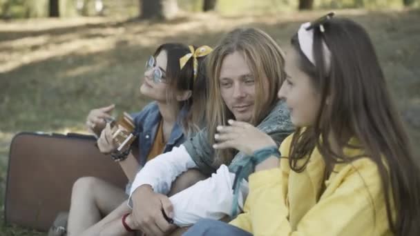 Side view of happy relaxed hippie friends sitting outdoors, chatting and playing ukulele. Positive young Caucasian 1960s man and women enjoying sunny day in summer park. Counterculture concept. — Stock Video