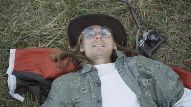 Close-up of relaxed hippie man in sunglasses lying on green grass and smiling at camera. Careless Caucasian 1960s guy enjoying relaxation outdoors on summer day. Pacifism and lifestyle. Stock Video