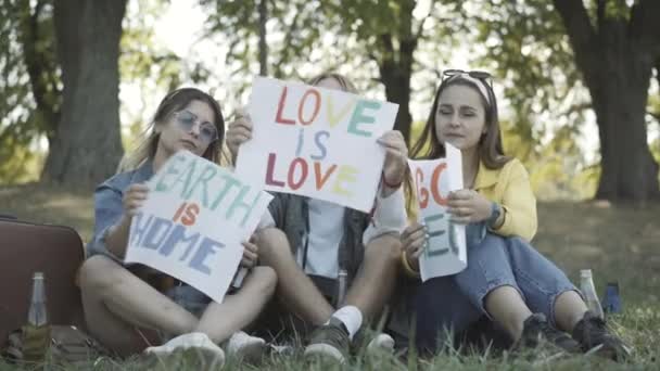 Drie jonge hippies tonen posters Aarde is thuis, Liefde is liefde, en Ga veganistisch, en kijkend naar de camera. Ontspannen blanke mannen en vrouwen die het pacifisme concept uitdrukken. Contra-cultuur van de jaren zestig. — Stockvideo