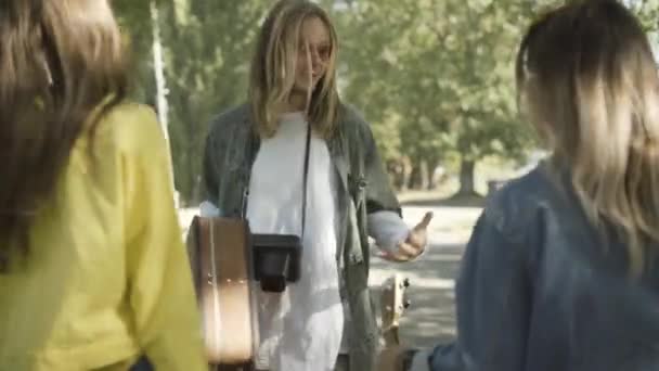 Joyful hippie man in sunglasses walking with suitcase and talking to women outdoors. Positive carefree 1960s Caucasian friends travelling on sunny summer day. Retro counterculture concept. — Stock Video