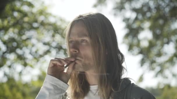 Close-up portrait of carefree 1960s hippie man smoking joint in sunlight outdoors. Young relaxed Caucasian man with long hair with cigarette on sunny summer day. Addiction and counterculture. — Stock Video