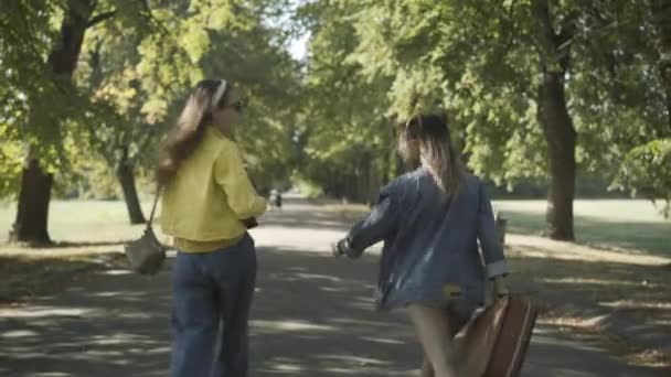 Vista posterior de dos mujeres hippies alegres caminando a lo largo de la carretera en el día soleado, girando a la cámara, y saludando lejos. Relajados despreocupados amigos jóvenes caucásicos que viajan en 1960 al aire libre. — Vídeos de Stock