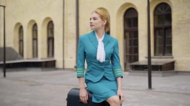 Şehir caddesinde seyahat çantasına oturmuş etrafa bakınan güzel, zayıf, kendine güvenen bir hostes. Açık havada bekleyen zarif, beyaz, sarışın bir kadının portresi. Seyahat ve zarafet konsepti.