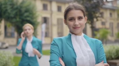 Kendine güvenen güzel hostes portresi kameraya bakıyor ve arka planda telefonda konuşan bulanık bir meslektaşı gibi gülümsüyor. Beyaz kadın şehir caddesinde poz veriyor..