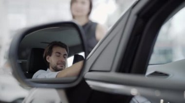 Arka planda konuşan bulanık kadını dinleyen gülümseyen yakışıklı adamın araba aynasındaki yansıması. Yeni bir araba galerisinde oturan mutlu bir beyaz müşteri. Otomobil endüstrisi.